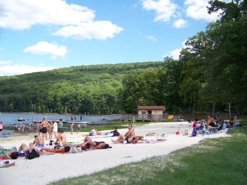 A sandy beach by a lake with people sunbathing, swimming, and enjoying the outdoors under a blue sky with clouds.