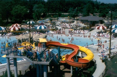A vibrant water park scene featuring a large slide, pools, and numerous sunbathers under colorful umbrellas.
