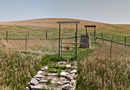 A grassy area with a gate leading to a historic dugout site, surrounded by a fence and open fields.