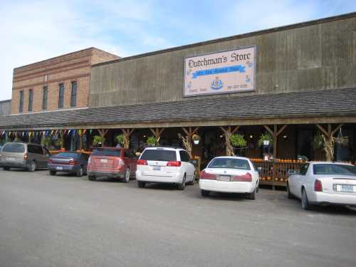 A rustic storefront with a sign reading "Dutchman's Store," featuring parked cars and hanging plants along the porch.