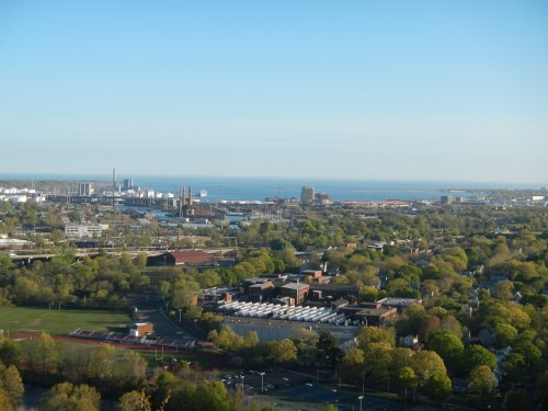 A panoramic view of a cityscape with greenery, industrial buildings, and a distant waterfront under a clear blue sky.