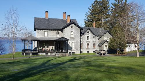 A large, historic stone house with a porch, surrounded by green grass and trees, near a body of water.