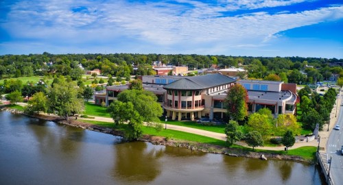 Aerial view of a modern building by a river, surrounded by green trees and a park under a blue sky.
