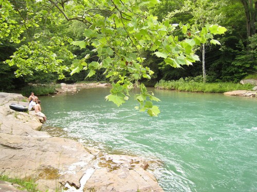 A serene river scene with people sitting on rocks, surrounded by lush green trees and clear turquoise water.