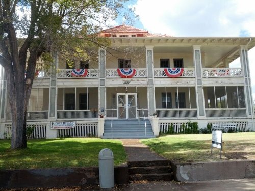 Historic building with a porch, decorated with red, white, and blue bunting, surrounded by greenery and a tree.