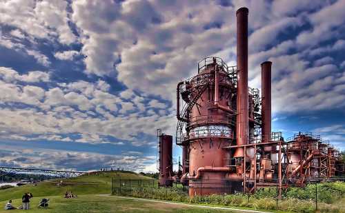 A large, rusted industrial structure with smokestacks, set against a cloudy sky and grassy area with people relaxing nearby.