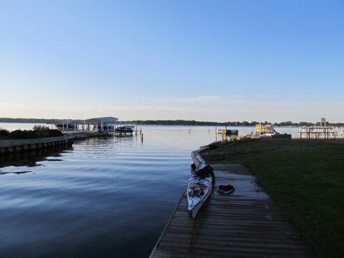 A calm lake scene with a wooden dock, a kayak, and distant buildings under a clear blue sky.