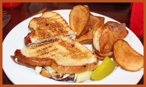 A grilled sandwich with melted cheese and a side of crispy potato chips and a pickle on a white plate.