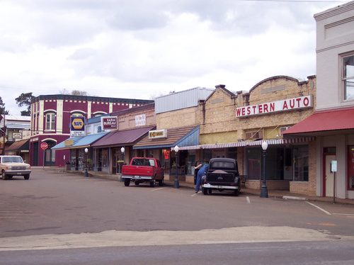 A small town street lined with vintage storefronts and parked cars under a cloudy sky.