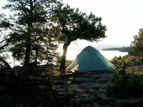 A tent set up near a lake, surrounded by trees, with sunlight reflecting on the water in the background.