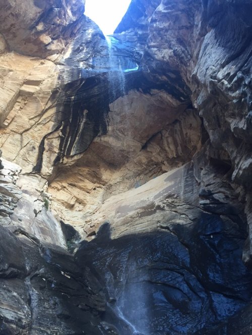 A narrow canyon with steep rock walls and a waterfall cascading down into the depths below, illuminated by sunlight.
