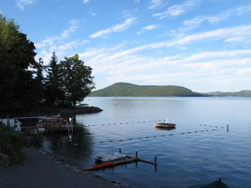 A serene lake scene with a dock, boats, and distant hills under a blue sky with scattered clouds.