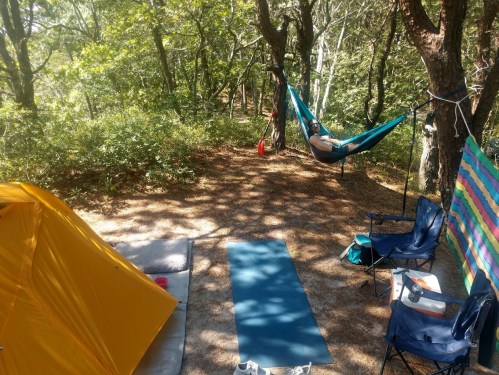 A cozy campsite with a yellow tent, hammock, blue yoga mat, and two camping chairs surrounded by trees.