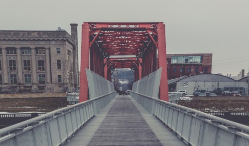 A red metal bridge spans a river, leading towards buildings in a gray, overcast setting.