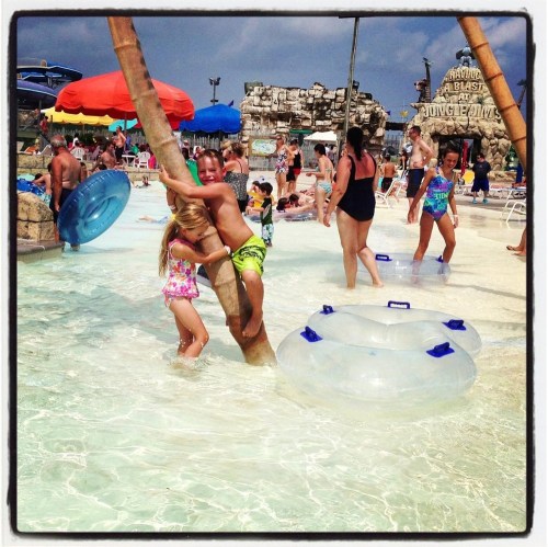 Children play in a water park, splashing in the pool and climbing on a palm tree, with colorful umbrellas in the background.