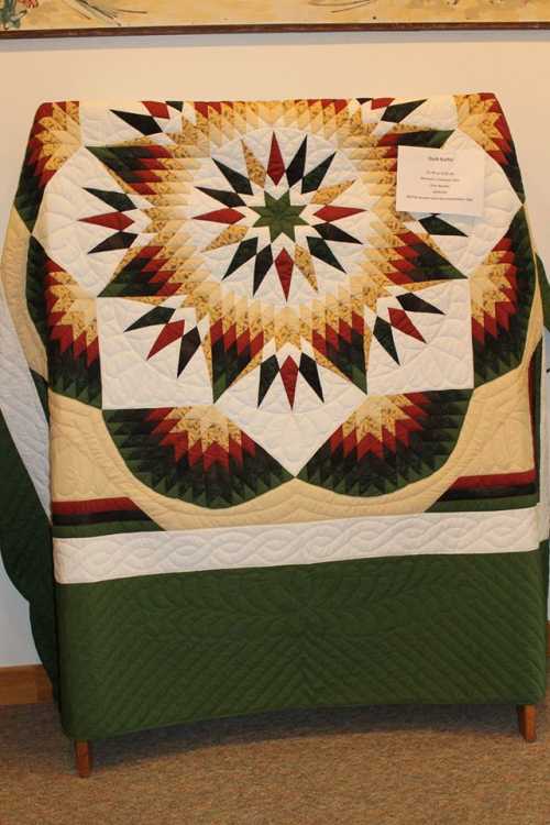 A colorful quilt displayed on a stand, featuring a star pattern with green, red, and cream colors.