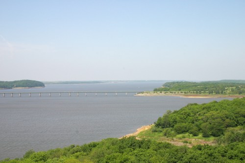 A serene view of a river with a bridge, surrounded by lush greenery and distant hills under a clear blue sky.
