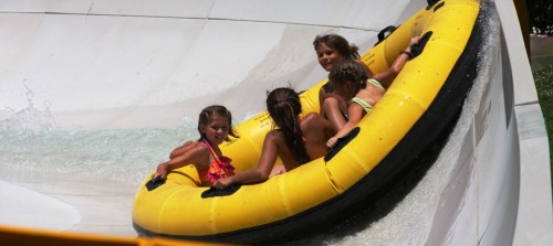 Four children enjoy a water slide in a yellow inflatable raft, splashing water around them.