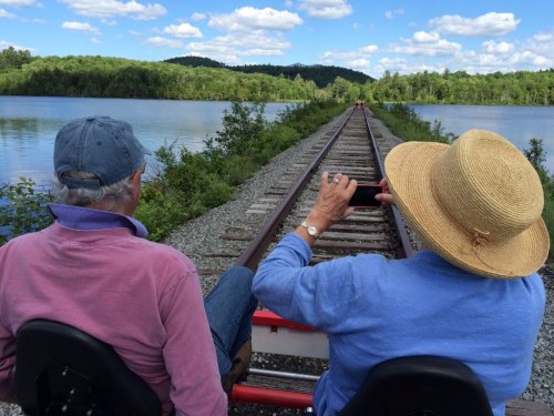 Two people sit on a rail bike, taking photos of a scenic view along a railway track by a lake and lush greenery.