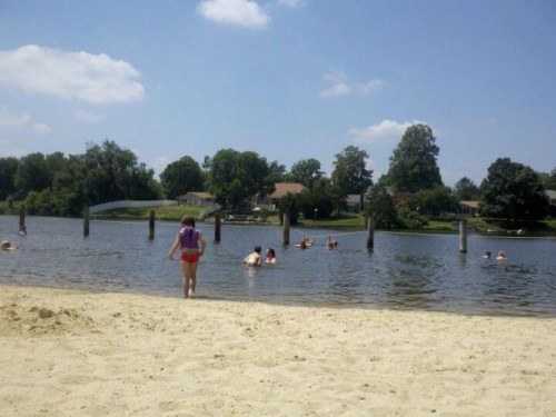 A sandy beach with a girl walking towards the water, where people are swimming and enjoying a sunny day.