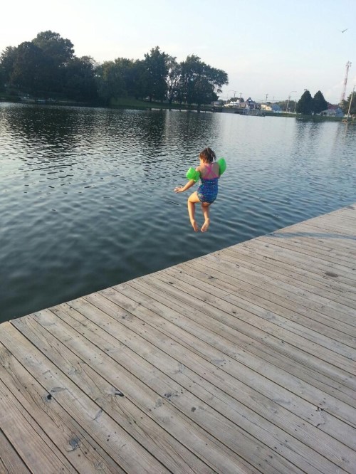A child in a swimsuit jumps off a wooden dock into a calm lake, wearing green floaties on their arms.