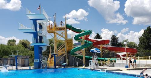 A vibrant swimming pool area featuring a diving board, water slides, and people enjoying the sunny day.