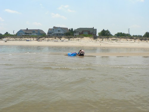 A person sits on a beach near the water, with houses visible in the background under a clear sky.