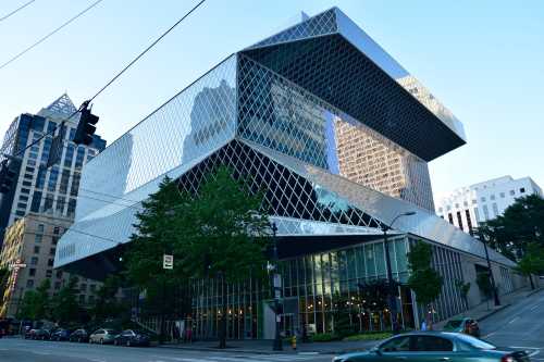 Modern glass building with a geometric design, surrounded by trees and city streets, reflecting the skyline.