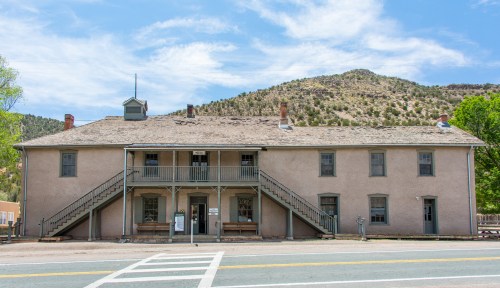 Historic building with two stories, stairs on both sides, set against a mountainous backdrop and blue sky.