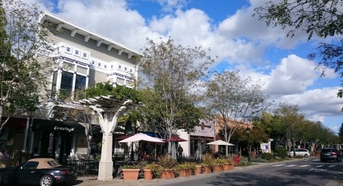 A charming street scene with shops, outdoor seating, and trees under a partly cloudy sky.