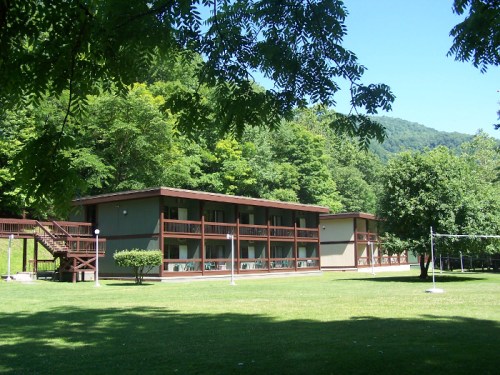 A two-story building surrounded by trees and a grassy area, with a volleyball net in the foreground.
