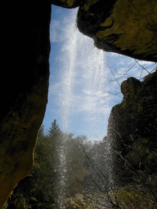 A waterfall cascades down from a rocky overhang, framed by trees and a blue sky in the background.