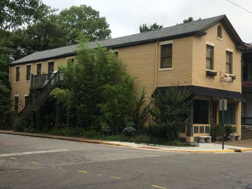 A two-story yellow building with a staircase, surrounded by greenery and a small garden, located at a street corner.