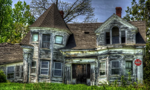 A weathered, abandoned house with a unique architectural design, featuring a turret and overgrown grass in the foreground.