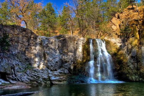 A serene waterfall cascades over rocky cliffs, surrounded by lush greenery and a clear blue sky.