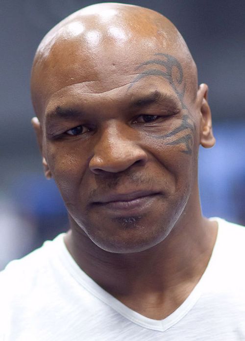 A close-up of a bald man with a tattoo on his face, wearing a white shirt and looking directly at the camera.