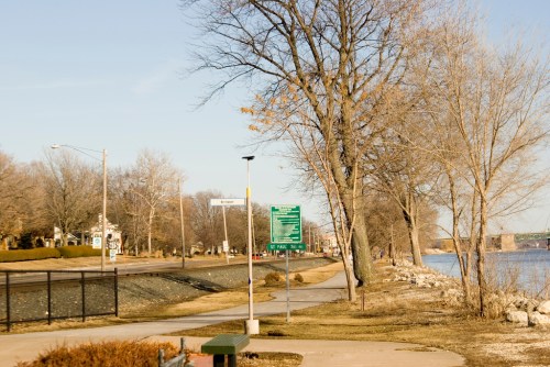 A scenic riverside path lined with trees, a signpost, and a view of the water on a clear day.