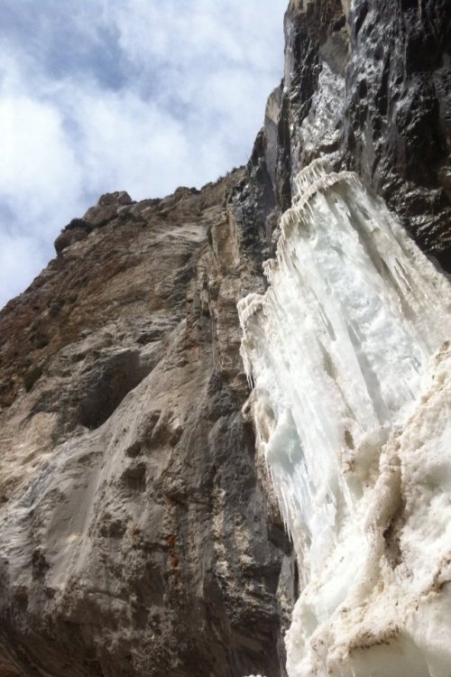 A towering ice formation clings to a rocky cliff under a cloudy sky.