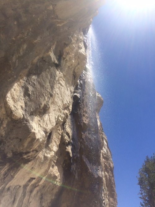 A waterfall cascading down a rocky cliff under a bright blue sky, with sunlight creating a misty spray.