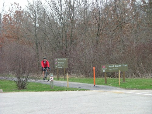 A cyclist in red rides on a path near signs for the Neal Smith Trail, surrounded by bare trees and grass.