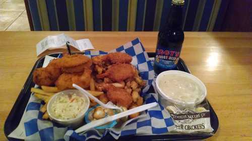 A tray with fried chicken, fries, coleslaw, a bowl of chowder, and a bottle of root beer on a wooden table.