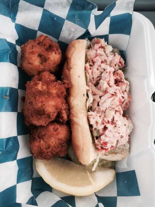 A sandwich filled with crab meat and coleslaw, served with two fried balls and a lemon wedge on a blue checkered paper.