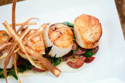 Three seared scallops on a bed of greens and sausage, garnished with crispy vegetable strips.