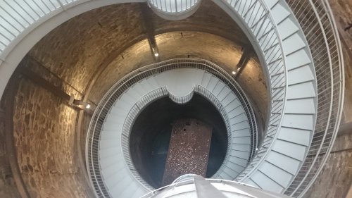 A spiral staircase inside a stone tower, viewed from above, leading down to a circular stone floor.