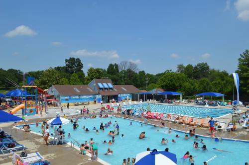 A busy outdoor pool area with people swimming, lounging, and enjoying the sun, surrounded by trees and blue skies.