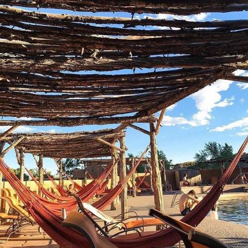 A serene outdoor space with hammocks hanging under a wooden structure, surrounded by blue skies and greenery.
