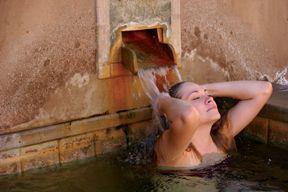 A woman relaxes in a warm bath, leaning back with water flowing from a stone spout behind her.