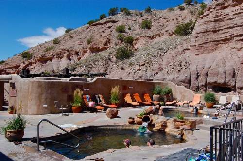 A serene hot spring pool surrounded by rocky terrain, with lounge chairs and people enjoying the water under a blue sky.