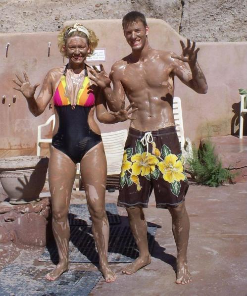 A man and woman covered in mud, smiling and posing with hands raised, wearing swimwear by a poolside.