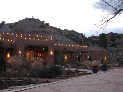 A rustic building with warm lights, surrounded by rocky terrain and sparse vegetation, under a cloudy sky.